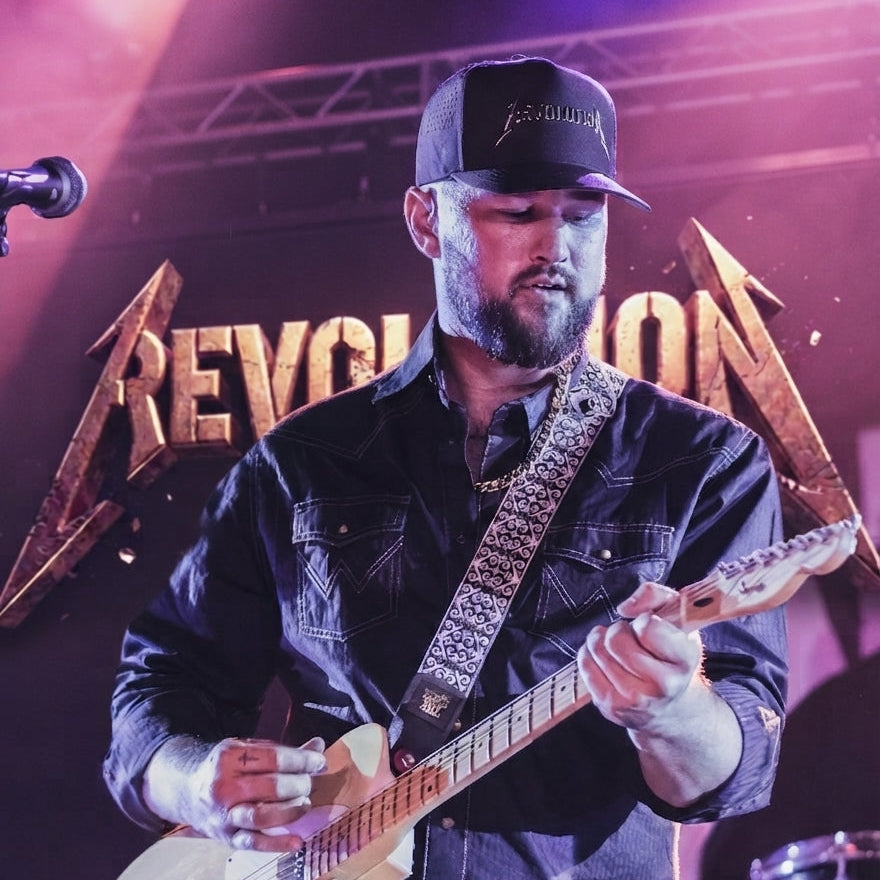 A male musician on stage playing a cream-colored electric guitar, wearing a black denim shirt, a black baseball cap, and a patterned guitar strap. The background features a large gold 'REVOLUTION' logo on a dark stage with purple and pink lighting.